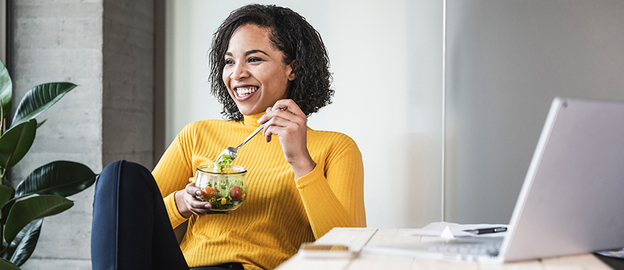 Person with black long hair wearing a yellow shirt sitting at their desk while smiling and eating a salad